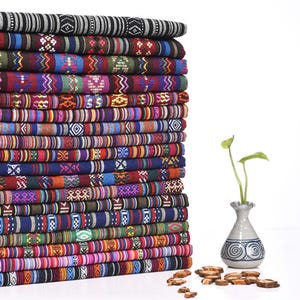 May include: A tall stack of folded fabric pieces featuring diverse woven patterns in vibrant colors including red, blue, green, purple, and black. Beside the stack sits a small ceramic vase with a green plant and scattered wood slices.