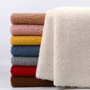Peut inclure: Une pile de tissu polaire sherpa plié dans diverses couleurs, notamment beige, rose clair, jaune, bleu, gris, rouge et marron foncé, avec un grand morceau de tissu polaire sherpa blanc drapé sur le côté de la pile de tissus colorés.