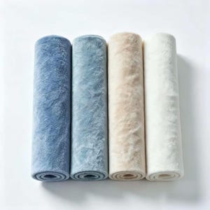 May include: Four cylindrical rolls of plush fabric arranged side by side. The colors from left to right are deep blue, light blue, beige, and off-white. Each roll features a soft, textured, fuzzy surface with a consistent pile throughout the length.