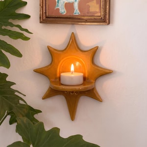 May include: A mustard yellow ceramic wall-mounted candle holder shaped like a sun with a central arched shelf. A small white tea light candle sits on the shelf with a lit flame, casting a warm glow against the glossy, star-pointed ceramic surface.
