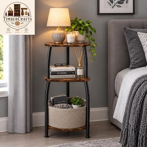 May include: A three-tier corner shelf with a dark wood finish and black metal frame. The top shelf holds a lamp and a candle. The middle shelf holds books titled The Kinfolk Home and At Home, and a reed diffuser. The bottom shelf holds a fabric basket.