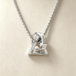May include: A silver-toned pendant necklace featuring a geometric triangular frame interlocked with a circular ring. The pendant hangs from a square-link chain. The metal surface displays a polished finish with subtle etched line patterns.