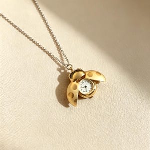 Peut inclure: Un collier pendentif doré en forme de coccinelle aux ailes ouvertes. Le centre de la coccinelle présente un petit cadran de montre analogique rond avec un fond blanc, des chiffres noirs et des aiguilles noires. Le pendentif est sur une chaîne argentée.