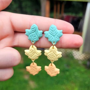 May include: A pair of dangle earrings featuring three stacked leaf-shaped charms in a gradient of colors. The top charms are teal, the middle are pale yellow, and the bottom are light orange. Each charm has a textured, embossed leaf vein pattern.