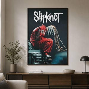 May include: A rectangular wall poster featuring the white text Slipknot at the top. Below the text, a figure in a red jumpsuit with a number 8 patch sits on a metal structure, wearing a textured grey mask with long, hanging dreadlock-like strands.