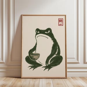 Könnte beinhalten: Ein gerahmter Wandkunstdruck, der einen dunkelgrünen Frosch mit weißem Bauch zeigt, der sitzt und eine kleine Schale hält. Der Frosch hat einen strengen Gesichtsausdruck. In der oberen rechten Ecke befindet sich ein roter Stempel mit den Zeichen 問一.