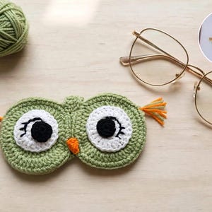 May include: A sage green crocheted owl-shaped eyeglass case featuring large white circular eyes with black pupils and eyelashes, a small orange beak, and orange yarn tufts on the sides. A ball of matching green yarn and gold-rimmed glasses are nearby.