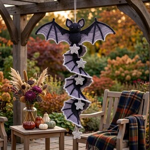 May include: A hanging crochet decoration featuring a dark purple and black bat at the top with a long, spiraling tail made of alternating purple and black segments. The spiral is accented with several small, white, five-pointed crochet stars.