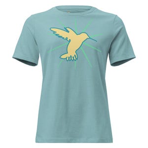 May include: A light blue short-sleeve t-shirt featuring a central graphic of a pale yellow hummingbird in flight. The bird is outlined in a dark teal color and is surrounded by radiating light green lines that extend outward from the center.