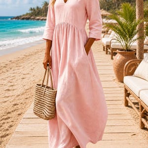 May include: A light pink maxi dress with a V-neckline, three-quarter length sleeves, and a gathered waistline featuring side pockets. The dress is paired with a woven straw tote bag and brown flat sandals, creating a simple and functional look.