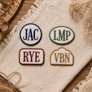 Puede incluir: Cuatro parches de tela bordados con fondos blancos y bordes de colores. Arriba a la izquierda, un parche circular con el texto azul marino JAC. Arriba a la derecha, un parche ovalado con texto verde LMP. Abajo a la izquierda, un parche rectangular con texto rojo RYE. Abajo a la derecha, un parche festoneado con texto dorado VBN.