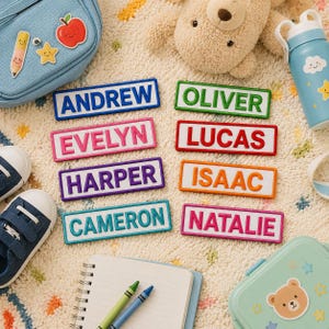 Op de afbeelding: Acht rechthoekige naamlabels met verhoogde letters in diverse kleuren, waaronder blauw, groen, roze, rood, paars, oranje, turquoise en magenta. De getoonde namen zijn Andrew, Oliver, Evelyn, Lucas, Harper, Isaac, Cameron en Natalie.