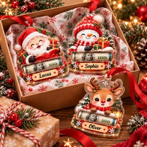 May include: Three clear acrylic Christmas ornaments featuring a Santa Claus, a snowman, and a reindeer. Each ornament includes a slot holding a rolled 100 dollar bill and a name tag reading Lucas, Sophie, or Oliver, finished with a red ribbon.
