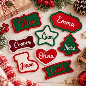 May include: A collection of felt name patches in various shapes including a bone, star, heart, Christmas tree, stocking, and rectangular labels. Each patch features embroidered names: Theo, Emma, Liam, Cooper, Nora, Jaxon, Olivia, and Caleb.