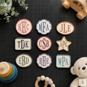 May include: Nine embroidered fabric patches featuring various shapes and monogrammed initials. The patches include circular, scalloped, oval, rectangular, and star designs with coloured borders and stitched letters in red, blue, orange, black, and green.