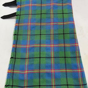Peut inclure: Un kilt avec un motif tartan dans des tons de vert et de bleu, rehaussé de fines lignes horizontales et verticales jaunes et rouges. Le vêtement comprend deux lanières en cuir noir avec des boucles métalliques fixées sur le côté pour le réglage.
