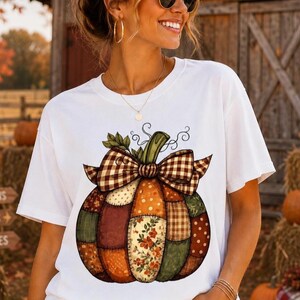 May include: A white short-sleeve t-shirt featuring a large graphic of a patchwork pumpkin. The pumpkin is composed of various fabric patterns including plaid, polka dots, and floral prints, topped with a brown and white checkered bow.