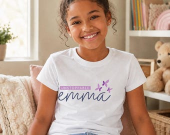 Peut inclure: Un t-shirt blanc à manches courtes avec le texte UNSTOPPABLE dans un rectangle violet au-dessus du prénom emma écrit en lettres cursives bleu foncé, accompagné de trois petits papillons violets sur le côté droit du texte.