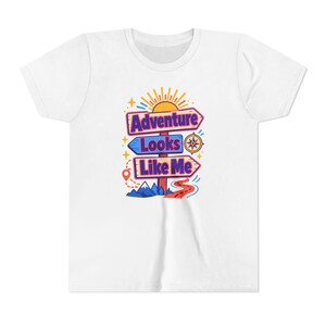 Peut inclure: Un t-shirt blanc à manches courtes avec un motif coloré représentant trois panneaux de signalisation en bois empilés portant le texte Adventure Looks Like Me. Le design inclut un soleil jaune, une boussole, des montagnes bleues et un chemin orange.