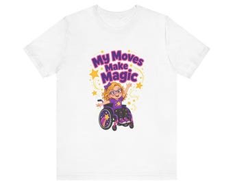 May include: A white short-sleeve t-shirt featuring a graphic of a person with blonde hair and glasses sitting in a wheelchair. The text above reads My Moves Make Magic in purple letters, surrounded by yellow stars and swirling lines.