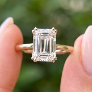 Pode incluir: Um anel com uma grande pedra preciosa transparente de corte esmeralda montada numa banda de ouro polido. A pedra retangular é fixada por quatro pequenas garras nos cantos, exibindo uma superfície facetada com um brilho transparente.