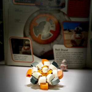 May include: A small, spherical construction toy assembled from white, orange, and dark gray plastic building blocks. The object features a central white stud surrounded by layered, curved segments. A tiny BB-8 droid figure stands in the background.