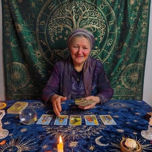 Pode incluir: Uma pessoa vestindo um top e um len&ccedil;o roxos senta-se a uma mesa coberta com um pano de veludo azul com padr&otilde;es celestiais dourados. Cinco cartas de tarot est&atilde;o alinhadas, com uma bola de cristal, uma vela acesa e um baralho por perto.