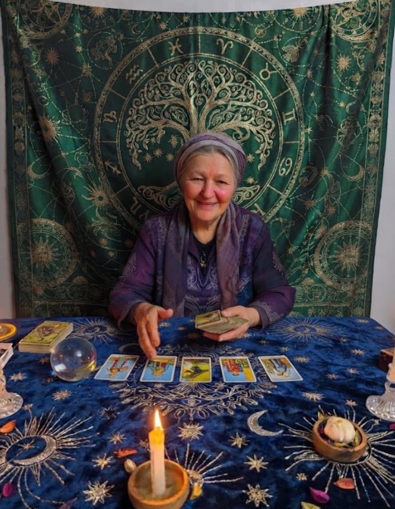 Pode incluir: Uma pessoa vestindo um top e um len&ccedil;o roxos senta-se a uma mesa coberta com um pano de veludo azul com padr&otilde;es celestiais dourados. Cinco cartas de tarot est&atilde;o alinhadas, com uma bola de cristal, uma vela acesa e um baralho por perto.