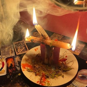 Pode incluir: Um prato de cer&acirc;mica branca cont&eacute;m duas velas cor-de-rosa verticais e uma vela bege horizontal, todas acesas com chamas e fumo vis&iacute;veis. O prato est&aacute; espalhado com folhas secas, agulhas de pinheiro e gotas de cera vermelha, rodeado de cartas.
