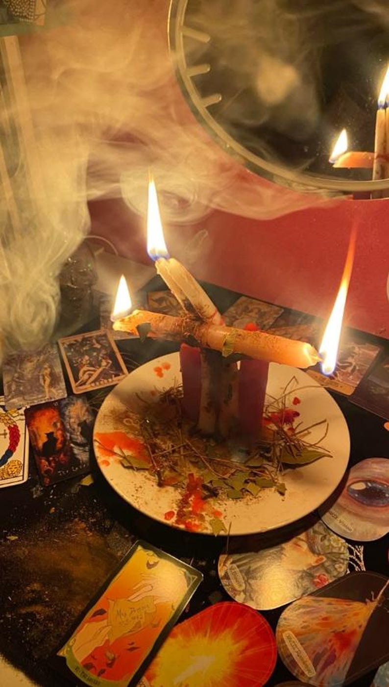 Pode incluir: Um prato de cer&acirc;mica branca cont&eacute;m duas velas cor-de-rosa verticais e uma vela bege horizontal, todas acesas com chamas e fumo vis&iacute;veis. O prato est&aacute; espalhado com folhas secas, agulhas de pinheiro e gotas de cera vermelha, rodeado de cartas.