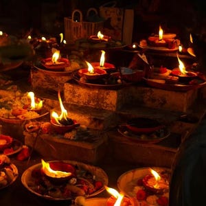Pode incluir: M&uacute;ltiplas pequenas l&acirc;mpadas de &oacute;leo de terracota com chamas tremeluzentes dispostas em degraus de pedra. Cada l&acirc;mpada assenta num prato contendo oferendas como p&eacute;talas de flores, bananas e outros pequenos itens, criando um brilho quente.