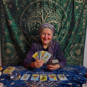 Pode incluir: Uma pessoa vestindo um top e um len&ccedil;o roxos segura um leque de quatro cartas de tar&ocirc;. Na mesa azul, cinco cartas de tar&ocirc; est&atilde;o dispostas em uma fileira ao lado de uma bola de cristal, um baralho e uma vela acesa. Uma tape&ccedil;aria verde com s&iacute;mbolos do zod&iacute;aco dourados e um design de &aacute;rvore est&aacute; pendurada ao fundo.