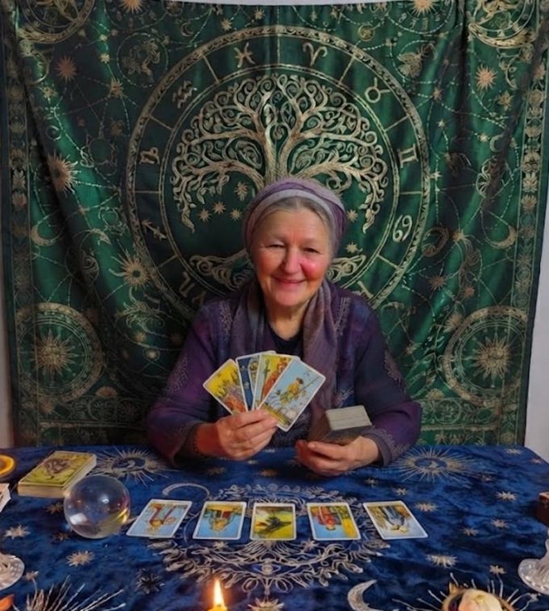 Pode incluir: Uma pessoa vestindo um top e um len&ccedil;o roxos segura um leque de quatro cartas de tar&ocirc;. Na mesa azul, cinco cartas de tar&ocirc; est&atilde;o dispostas em uma fileira ao lado de uma bola de cristal, um baralho e uma vela acesa. Uma tape&ccedil;aria verde com s&iacute;mbolos do zod&iacute;aco dourados e um design de &aacute;rvore est&aacute; pendurada ao fundo.