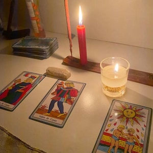 Pode incluir: Uma tiragem de cartas de tarot com as cartas Valete de Copas e XIX-O Sol, acompanhada por uma vela vermelha acesa num suporte de madeira, uma pequena vela votiva branca num frasco de vidro, um baralho de cartas e uma pequena pedra clara.