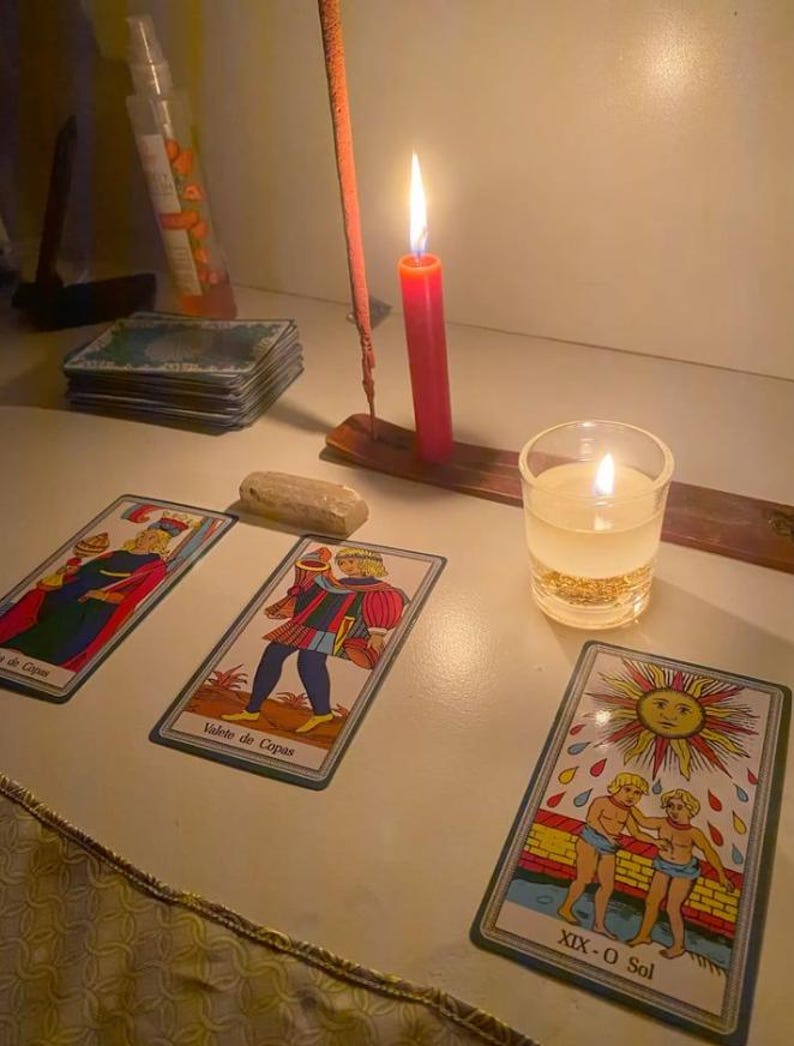 Pode incluir: Uma tiragem de cartas de tarot com as cartas Valete de Copas e XIX-O Sol, acompanhada por uma vela vermelha acesa num suporte de madeira, uma pequena vela votiva branca num frasco de vidro, um baralho de cartas e uma pequena pedra clara.