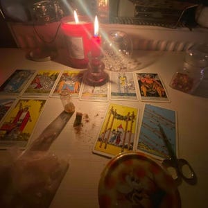 Pode incluir: Uma cole&ccedil;&atilde;o de cartas de tar&ocirc; dispostas sobre uma superf&iacute;cie com duas velas vermelhas acesas, um pequeno frasco de vidro, uma tesoura de metal e um recipiente redondo com uma ilustra&ccedil;&atilde;o de abelha, iluminados pelo brilho das velas.