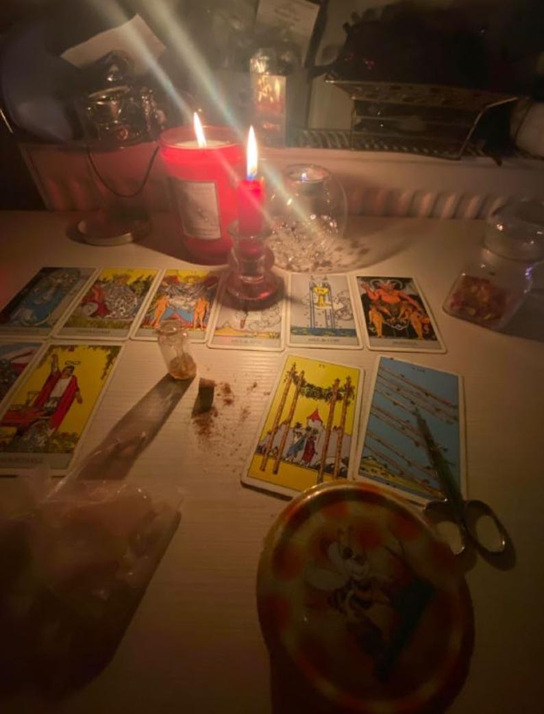 Pode incluir: Uma cole&ccedil;&atilde;o de cartas de tar&ocirc; dispostas sobre uma superf&iacute;cie com duas velas vermelhas acesas, um pequeno frasco de vidro, uma tesoura de metal e um recipiente redondo com uma ilustra&ccedil;&atilde;o de abelha, iluminados pelo brilho das velas.