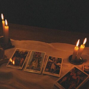 Pode incluir: Quatro cartas de tarot est&atilde;o dispostas sobre um pano branco, com uma carta exibindo o texto O DIABO. As cartas s&atilde;o iluminadas por quatro velas acesas em suportes de metal, projetando um brilho quente sobre um fundo escuro.