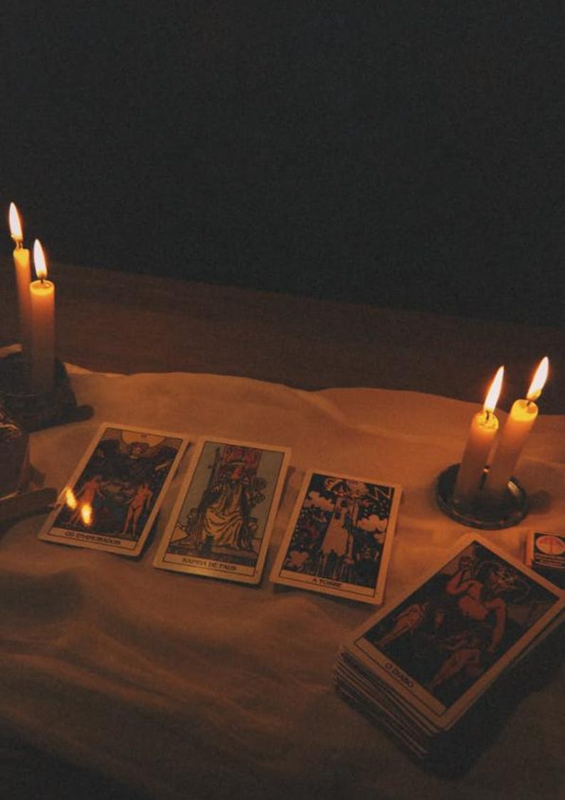 Pode incluir: Quatro cartas de tarot est&atilde;o dispostas sobre um pano branco, com uma carta exibindo o texto O DIABO. As cartas s&atilde;o iluminadas por quatro velas acesas em suportes de metal, projetando um brilho quente sobre um fundo escuro.