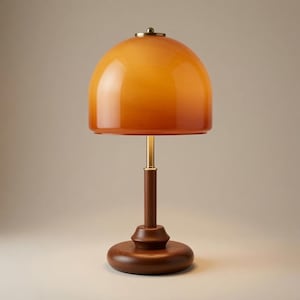 Peut inclure: Une lampe de table dotée d'un abat-jour en verre orange en forme de dôme lisse avec un dégradé subtil. La lampe possède un col en métal couleur laiton fin et une base circulaire en bois brun foncé qui assure la stabilité de l'ensemble.