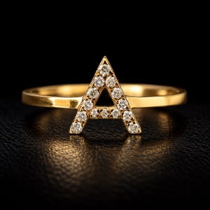 May include: A gold ring featuring a capital letter A as the centrepiece. The letter A is encrusted with small, clear, round-cut stones that sparkle against the polished gold band. The design is simple, with the letter integrated into the ring band.