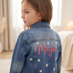 May include: A blue denim jacket featuring a back panel embroidered with the name Mazie in pink script lettering, surrounded by multiple small white daisy flower motifs with yellow centres, including a classic collar and long sleeves.