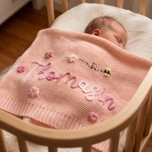 May include: A soft, light pink knitted baby blanket featuring the embroidered name Thomasin in a darker pink script. The blanket is decorated with small, three-dimensional floral accents and a small embroidered bee with a dotted flight path.