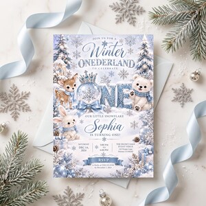 May include: A rectangular invitation card featuring a Winter Onederland theme with a fawn, a rabbit, and a polar bear wearing blue scarves. The text reads Winter Onederland, Sophia is turning one, and includes event details for December 14, 2024.