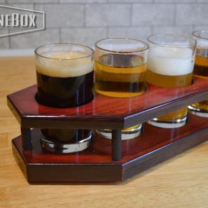 Puede incluir: Una tabla de degustación de cerveza de madera oscura con forma de ataúd, que presenta cuatro aberturas circulares con cuatro vasos de vidrio transparente llenos de diversas cervezas. La palabra PINEBOX está impresa en blanco en la esquina.