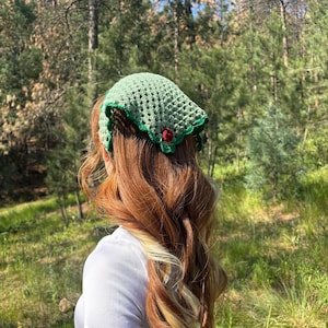 May include: A sage green triangular crochet headscarf featuring a delicate openwork stitch pattern, a dark green scalloped border, and a small red crocheted strawberry accent attached to the bottom point of the fabric.
