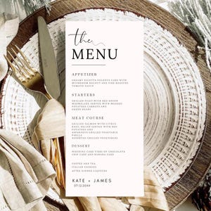 May include: A white rectangular wedding menu card with black text titled The Menu, listing courses including appetizer, starters, meat course, and dessert, ending with the names Kate + James and the date 07.12.20XX, placed on a decorative plate.