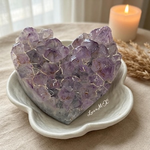 Puede incluir: Un racimo de cristales de amatista en forma de corazón con puntas facetadas de color púrpura intenso. El cristal descansa dentro de un plato de cerámica blanco con bordes festoneados. El texto "LuceMX" está impreso en cursiva blanca.