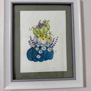 Puede incluir: Una pintura en acuarela enmarcada que muestra una pila de tres calabazas en tonos verde, blanco y azul. Las calabazas están decoradas con flores moradas, girasoles amarillos y margaritas blancas pintadas, todo dentro de un marco blanco y morado.