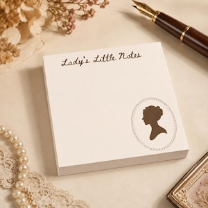 May include: A square notepad with cream-colored paper featuring the brown text Lady's Little Notes at the top and a brown silhouette of a person's profile inside an oval decorative frame in the bottom right corner of the top sheet.